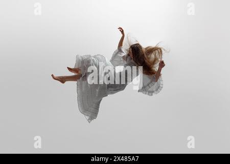 Woman dressed in flowing white gown suspend in mid-air, her long blonde hair cascading around her against white studio background. Stock Photo