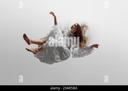 Young woman, dressed in elegant white dress with delicate ruffles, seems to levitate against white studio background. Stock Photo