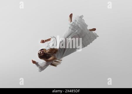 Elegantly dressed in white gown, woman floats, falling down in mid-air in delicate pose, against white studio background. Stock Photo