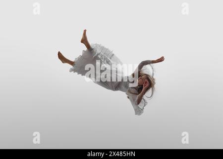 Elegantly dressed in white vintage gown, woman floats, falling down in mid-air in delicate pose, against white studio background. Stock Photo