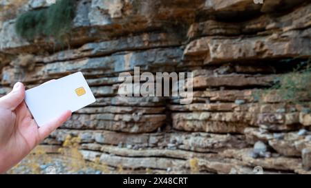 In a woman's hand, a white bank card against a brownish-red rocky cliff background.  Stock Photo