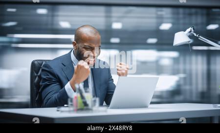 Office: Happy Successful Black Businessman Sitting at Desk Using Laptop Computer, Celebrate Success. Entrepreneur in Suit working with Stock Market App Smiles, Happy Victory. Motion Blur Background. Stock Photo