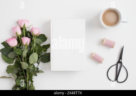 White book mockup with a bouquet of pink roses and workspace accessories on a white table. Stock Photo