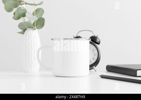 Enamel mug mockup with eucalyptus in a vase and workspace accessories on a white table. Stock Photo