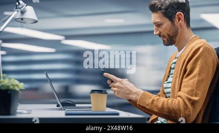 Modern Office: Businessman Sitting at His Desk Using Smartphone, Laptop Computer. Digital Entrepreneur working with Big Digital Data e-Commerce. Motion Blur Background Showing Active Work Day. Stock Photo