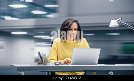Modern Office: Happy Smiling Hispanic Businesswoman Sitting at Her Desk Working on a Laptop Computer Celebrates Victory. Latin Female Entrepreneur is a Happy Winner. Motion Blur Background Stock Photo
