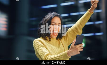 Street Shot: Portrait of Beautiful Latin Woman Using Smartphone, Celebrating Successful Victory with Yes Gesture. Smiling Hispanic Female Entrepreneur Using Mobile Phone Happily. Blur Motion Shot. Stock Photo