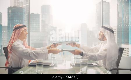 Two Successful Emirati Businessmen in White Traditional Kandura Sitting in Office and Signing Contract. Business Partners Exchange Documents on Lucrative Investment. Saudi, Arab Businessman Concept. Stock Photo