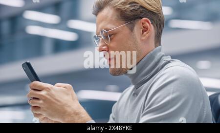 Modern Office: Portrait of Creative Businessman Sitting at His Desk Using Smartphone. Digital Entrepreneur Using Mobile Phone, Internet e-Commerce Online Shopping. Blurred Background. Stock Photo