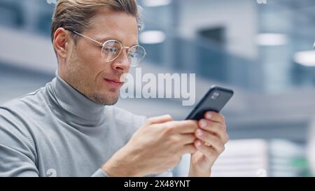 Modern Office: Portrait of Creative Businessman Sitting at His Desk Using Smartphone. Digital Entrepreneur Using Mobile Phone, Internet e-Commerce Online Shopping. Blurred Background. Stock Photo