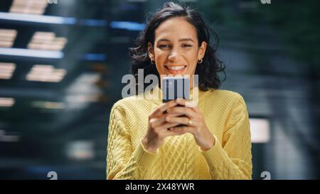 Street Shot: Portrait of Beautiful Latin Woman Using Smartphone. Smiling Hispanic Female Entrepreneur Using Mobile Phone for Online Shopping, e-Commerce Happily. Blurred Motion Background. Stock Photo