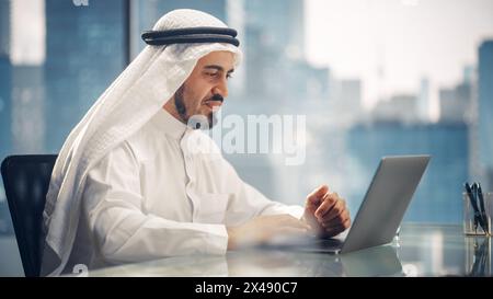 Arab Businessman in White Traditional Outfit Sitting in Office and Working on Laptop Computer. Business Manager Make Successful Investment Deal. Saudi, Emirati, Arab Businessman Concept. Stock Photo