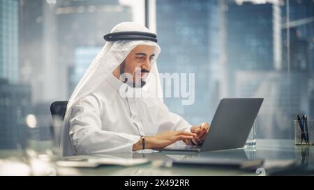 Arab Businessman in White Traditional Outfit Sitting in Office and Working on Laptop Computer. Business Manager Make Successful Investment Deal. Saudi, Emirati, Arab Businessman Concept. Stock Photo