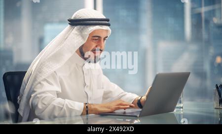 Arab Businessman in White Traditional Outfit Sitting in Office and Working on Laptop Computer. Business Manager Make Successful Investment Deal. Saudi, Emirati, Arab Businessman Concept. Stock Photo