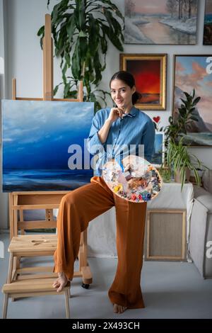 Portrait artist woman stands near easel with canvas in art studio hold colour palette look at camera Stock Photo