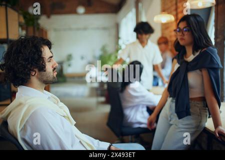 Young people, freelancers occupying a shared workspace, with someone focused on digital screens and others engaged in active discussions. Stock Photo