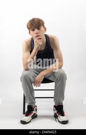 A Fifteen Year Old Teenage Boy in a sleeveless top and tracksuit bottoms sitting in A Chair leaning forward Stock Photo