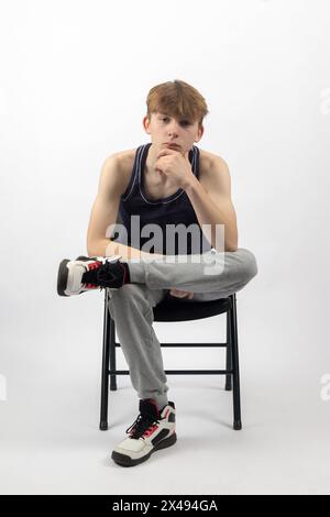 A Fifteen Year Old Teenage Boy in a sleeveless top and tracksuit bottoms sitting in A Chair leaning forward Stock Photo