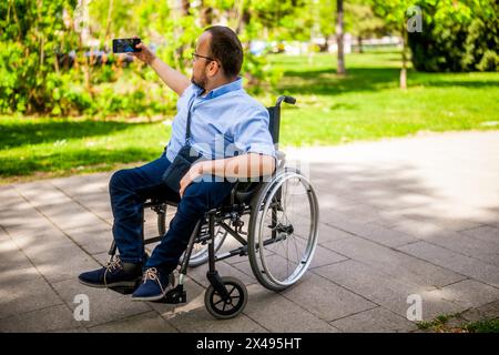 Portrait of happy man in wheelchair. He is enjoying sunny day in city park and taking selfie. Stock Photo