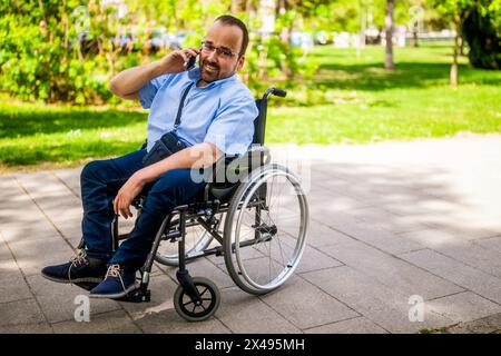 Portrait of happy man in wheelchair. He is enjoying sunny day in city park and talking on phone. Stock Photo