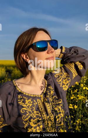 Ukrainian woman in shirt embroidered with yellow ornament among field of blooming rapeseed flowers. Independence Day, Vyshivanka Day. Ukrainians ask f Stock Photo