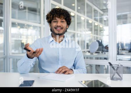 Indian male spreading hands with pen to camera while sitting in front of table with notebook. Smiling hr manager asking question from candidates for hiring and making remarks in copybook. Stock Photo