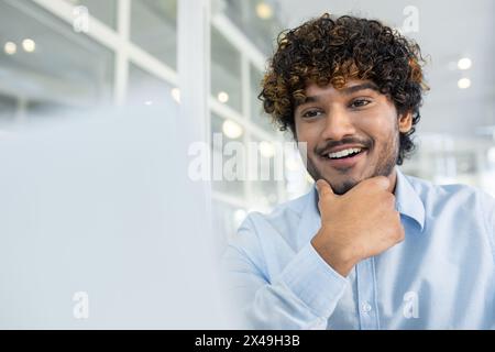 Selective focus on arab trainee holding chin with hand while looking excited at screen of computer. Enthusiastic junior specialist watching webinar and learning new material for work indoors. Stock Photo