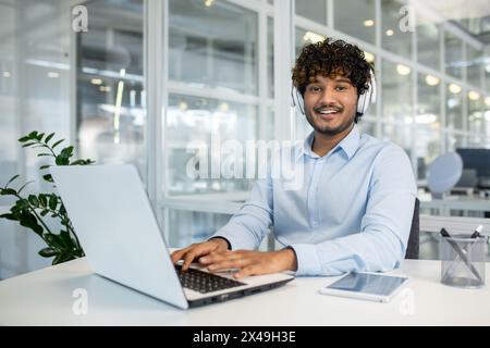 Member of client's support team wearing white headphones and using portable computer in modern workplace. Middle eastern guy listening audio complains and getting ready offering problem solutions. Stock Photo
