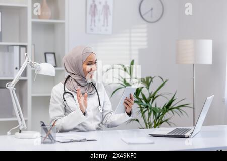 Arabic female doctor in hijab greeting remote client while providing online consultation from private clinic. Medic professional sitting by white desk with laptop and using digital tablet in cabinet. Stock Photo