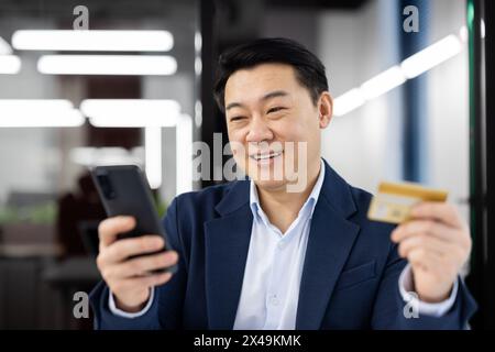 Positive middle age man using modern smartphone and holding plastic bank card on blurred background. Friendly economy specialist helping client connecting payment accessibility with application. Stock Photo