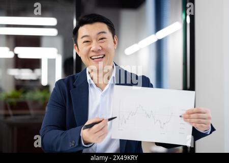 Cheerful taiwan investor showing sheet of paper with growing statistics and pointing with pen to necessary spot. Financial expert felling proud about result of work and made decisions in trading. Stock Photo
