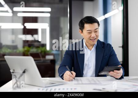 Smiling chinese accountant summarising expenses and income while using calculator and writing down indicators. Attentive economist filling document wi Stock Photo