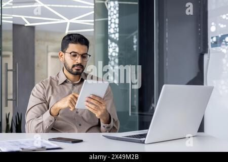 Concentrated hispanic freelancer checking list in notebook while leaning on chair by desktop with pc. Young professional reading remarks written in diary while sitting in stylish office interior. Stock Photo