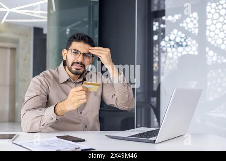 Hopeless middle eastern male touching forehead with hand while looking at camera and holding credit card in hand. Young guy trying to accomplish online purchase and feeling frustrated with process. Stock Photo