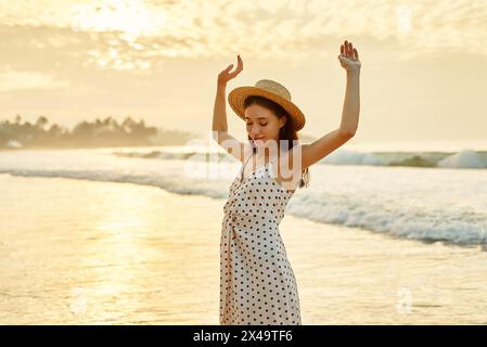 Elegant lady in polka dot dress enjoys sunset on beach with arms raised. Solo traveler feels free by sea embodies summer leisure escape. Warm Stock Photo Alamy