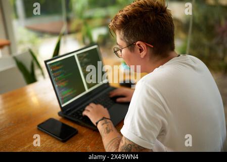 Transgender web developer works on laptop, programming in modern eco office. Focused individual with tattoos types code, designs app. Commitment to Stock Photo