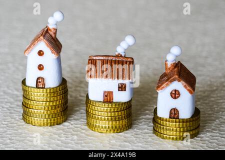Three different small toy houses on yellow coin stacks of different heights on grey background, different cost concept Stock Photo