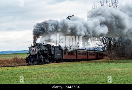 Ronks, Pennsylvania, USA, December 30, 2023 - A steam train is traveling down the tracks, leaving a trail of smoke behind it. The train is surrounded Stock Photo