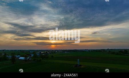 Aerial View of Dramatic Sunset Casting A Warm Glow Over A Rural Landscape With Silhouetted Trees And Buildings Under A Cloud-Streaked Sky. Stock Photo