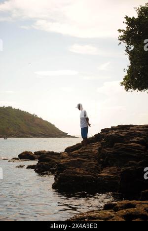 a moment of serenity by the ocean a lone man standing on the rugged rocky shore gazing into the vast sea with an island on the horizon Stock Photo