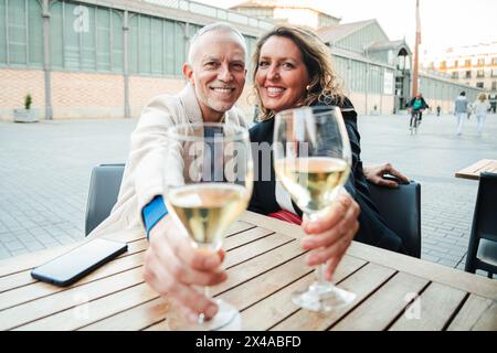 Mature smiling couple toasting white wine glasses to the camera. Middle aged married people drinking alcohol on a bar terrace. Two caucasian adult partners clinking with alcohol beverages at vacations. High quality photo Stock Photo
