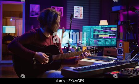 Portrait of male songwriter playing acoustic guitar and recording musical sounds to use for mix and master in home studio. Composing tunes with digital audio workstation software. Camera B. Stock Photo