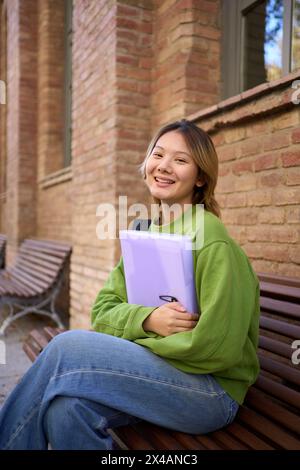 Portrait of young smiling Chinese student sitting on a bench on campus looking at the camera Stock Photo