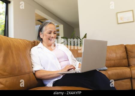 Asian senior woman using laptop at home, sitting on a couch. Wearing a white shirt and black pants, she has short gray hair, unaltered Stock Photo