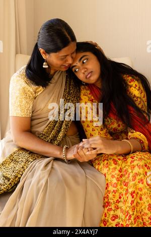 Indian mother and teenage daughter, holding hands in cozy living room. Both with long black hair, dressed in yellow and beige, sharing smiles and feel Stock Photo