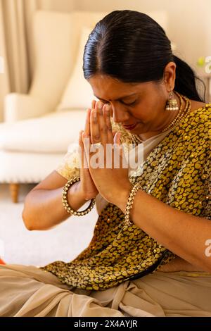 Indian woman sitting at home, hands pressing together, eyes closing. Wearing gold jewelry, with long black hair and mature skin, she exudes calmness a Stock Photo