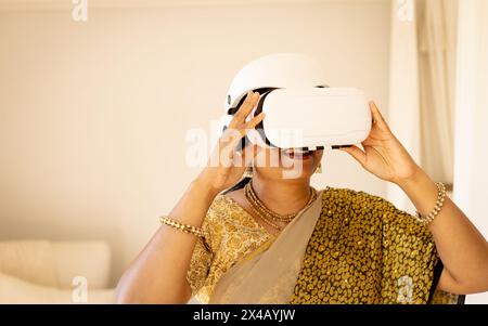Indian woman wearing virtual reality headset at home, exploring new tech. Dressed in traditional attire, with dark hair and gold jewelry, enjoying imm Stock Photo