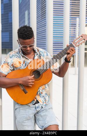 Stylish young man focused on playing his acoustic guitar outdoors, dressed in a colorful tropical shirt. Stock Photo