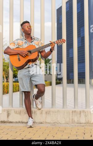 Vertical image of a fashionable young African man playing guitar in an urban park, featuring stylish summer wear and architectural background. Stock Photo