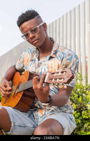 Close-up view of a young African man with stylish glasses and a colorful shirt, deeply focused on playing an acoustic guitar outdoors. Stock Photo